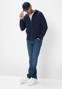 Felpa con zip di colore navy, dal design aderente, indossata sopra una t-shirt bianca, abbinata a jeans blu e sneakers bianche; presenta un cappuccio e un tessuto elegante.