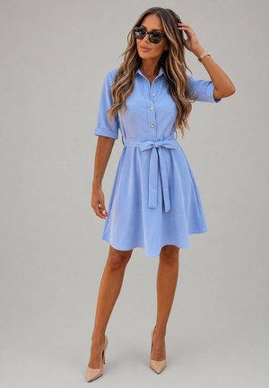 Femme en robe-chemise bleu clair jusqu'aux genoux avec ceinture à la taille, portant des lunettes de soleil et des talons hauts couleur nude, debout devant un fond uni.