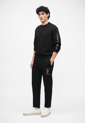 Sweatshirt preto com texto dourado na manga, combinado com calças de treino pretas com um gráfico dourado. Os sapatos são brancos com uma sola texturizada.
