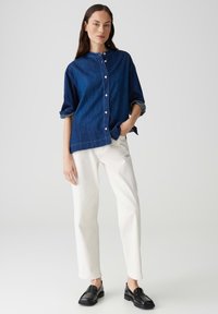 Camicia button-up in jeans di colore blu scuro, con colletto rotondo e maniche arrotolate. Abbinata a pantaloni bianchi a gamba dritta e mocassini neri.