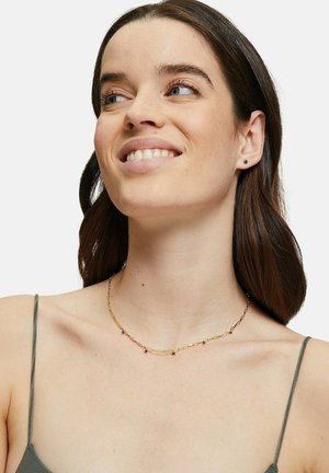 Collier en chaîne dorée avec des maillons allongés et de petits accents en pierre bleue, présenté sur un mannequin avec un fond neutre.