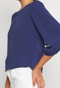 Blouse bleu marine à la coupe décontractée, manches bouffantes trois-quarts, lueur subtile et encolure arrondie. Ourlet droit avec fentes latérales.