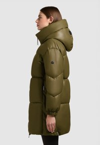 Luchtig groene parka met een hoge nek en capuchon, voorzien van een gladde textuur, elastische manchetten en een groot zwart embleem op de mouw.