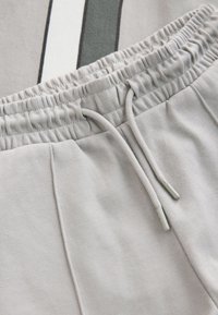 Vaaleanharmaat collegehousut, joissa on joustava vyötärönauha ja kiristysnauha sekä kaksi pystysuoraa sivupaneelia tummemmissa sävyissä. Pehmeä kangas.