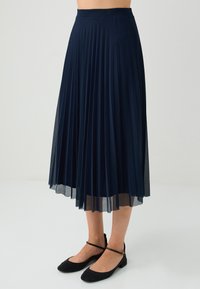 Gonna midi plissettata blu navy con elastico morbido in vita e tessuto a strati semi-trasparente. Abbinata a ballerine nere, che mostrano un design minimalista.