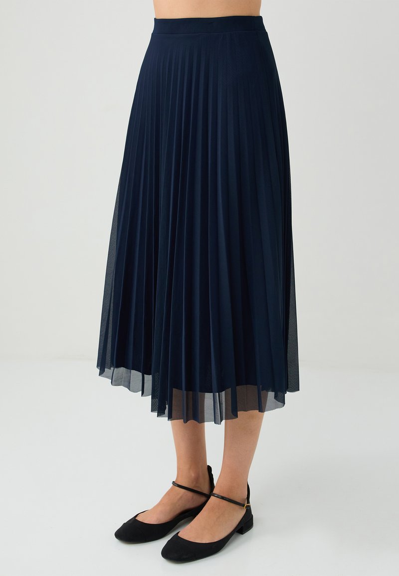 Gonna midi plissettata blu navy con elastico morbido in vita e tessuto a strati semi-trasparente. Abbinata a ballerine nere, che mostrano un design minimalista.