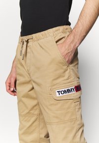 Khaki spodnie cargo z elastycznym pasem i sznurkiem, z bocznymi kieszeniami i naszywką z napisem "TOMMY JEAN" w kolorach czerwonym, białym i niebieskim.