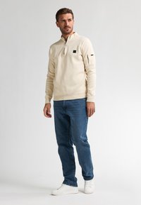 Pullover in pile color avorio da uomo con mezza zip, patch logo nero sul petto e tasca laterale; abbinato a jeans blu e sneakers bianche.