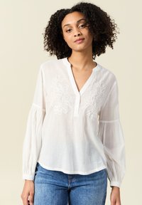 Blusa di cotone bianca con scollo a V, decorazioni floreali ricamate e maniche lunghe e ampie. Indossata con jeans blu di denim.