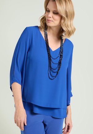 Kvinde iført en lagdelt royalblå bluse og bukser med en multilags mørkeblå perlekæde, der kigger ned og til siden.