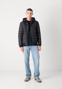 Sort pufferjakke med hætte, med quiltet design og lynlås lukning. Parret med en marineblå skjorte, lyseblå jeans og grå sko.
