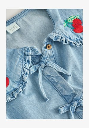 Világoskék farmer póló, fodros gallérral, fából készült gombbal és cseresznye hímzett díszítéssel. Puha tapintású, megkötős záródással.