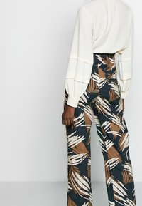 Vit långärmad topp med lager av trumpetärmar, kombinerad med högmidjade, marinblå byxor med ett brunt och vitt lövmönster.