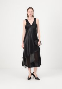 Black dress featuring a deep V-neck and an asymmetrical layered design. Made of sheer and satin-like material. Accessorized with a small black bag.