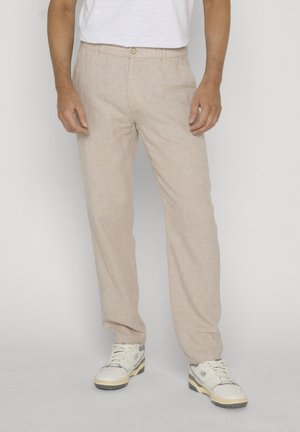 Mężczyzna w beżowych lnianych spodniach i białych trampkach, stoi na jednolitym jasnoszarym tle.