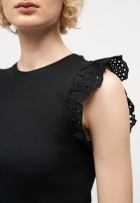 Γυναίκα που φοράει μαύρο αμάνικο τοπ με δαντελένιες λεπτομέρειες βολάν στους ώμους, μπροστά σε απλό ανοιχτό φόντο.