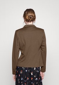 Mujer con el cabello castaño claro recogido en un moño, que lleva pendientes de aro dorados, un blazer marrón ajustado y una falda negra con flores, de espaldas frente a un fondo neutro.