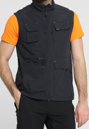 Hombre con chaleco utilitario negro de múltiples bolsillos sobre camisa de manga corta de color naranja brillante y pantalones negros, con las manos parcialmente en los bolsillos.