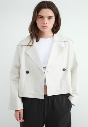 Kobieta o długich brązowych włosach nosi krótką kremową dwurzędową kurtkę na białym topie i luźnych czarnych spodniach, stoi na jednolitym białym tle.