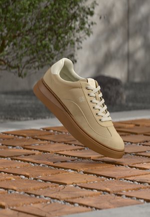 Baskets en daim beige avec lacets blancs et semelle marron, flottant au-dessus d'un pont en bois, plante verte et mur gris en arrière-plan.