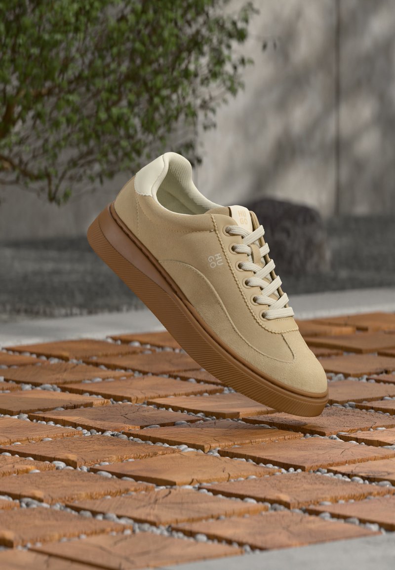 Μπεζ σουέντ αθλητικό παπούτσι με λευκά κορδόνια και καφέ σόλα που αιωρείται πάνω από ξύλινο δάπεδο, με πράσινο φυτό και γκρί τοίχο στο φόντο.