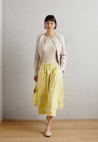 Veste en cuir beige, top crème, jupe midi à fleurs jaunes et chaussures crème. Porte un sac à main jaune. Fond neutre et sol en bois.