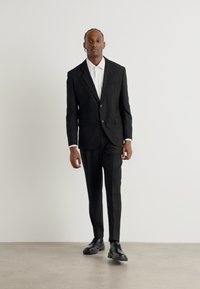 Schwarzer Anzug mit taillierter Jacke und schmalen Hosen, kombiniert mit einem weißen Hemd. Glattes Material, minimalistisches Design, getragen mit schwarzen Schnürschuhen.