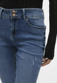 Los pantalones vaqueros de mezclilla oscura presentan una cintura alta con cierre de doble botón, bolsillos laterales y ligeros detalles desgastados en la parte frontal.
