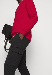 Hombre con camisa de manga larga roja y pantalones cargo negros sosteniendo una bolsa negra con correas verdes frente a un fondo simple.