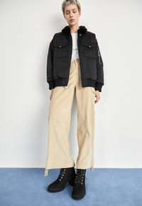 Black bomber jacket with fur collar, beige wide-leg corduroy pants, and black lace-up ankle boots. Simple design, textured materials.