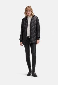 Giacca imbottita nera trapuntata con cappuccio, dotata di cerniera frontale e tasche laterali, indossata sopra a un maglione bianco e abbinata a pantaloni di pelle neri.