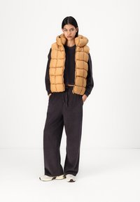 Gilet imbottito senza maniche color miele con cappuccio, abbinato a un maglione lavorato a maglia scuro e pantaloni a gamba larga in velluto a coste. Le scarpe sono sneakers beige chiaro.