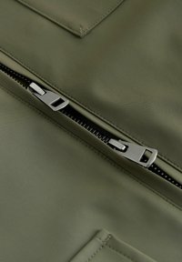 Sølvmetal lynlås delvist åbnet på mørkegrønt stof med synlige syninger og en rektangulær lommeflap.