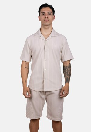 Hombre con pelo oscuro vistiendo una camisa beige de manga corta con botones y pantalones cortos a juego, de pie con expresión neutral y tatuajes visibles en los brazos.