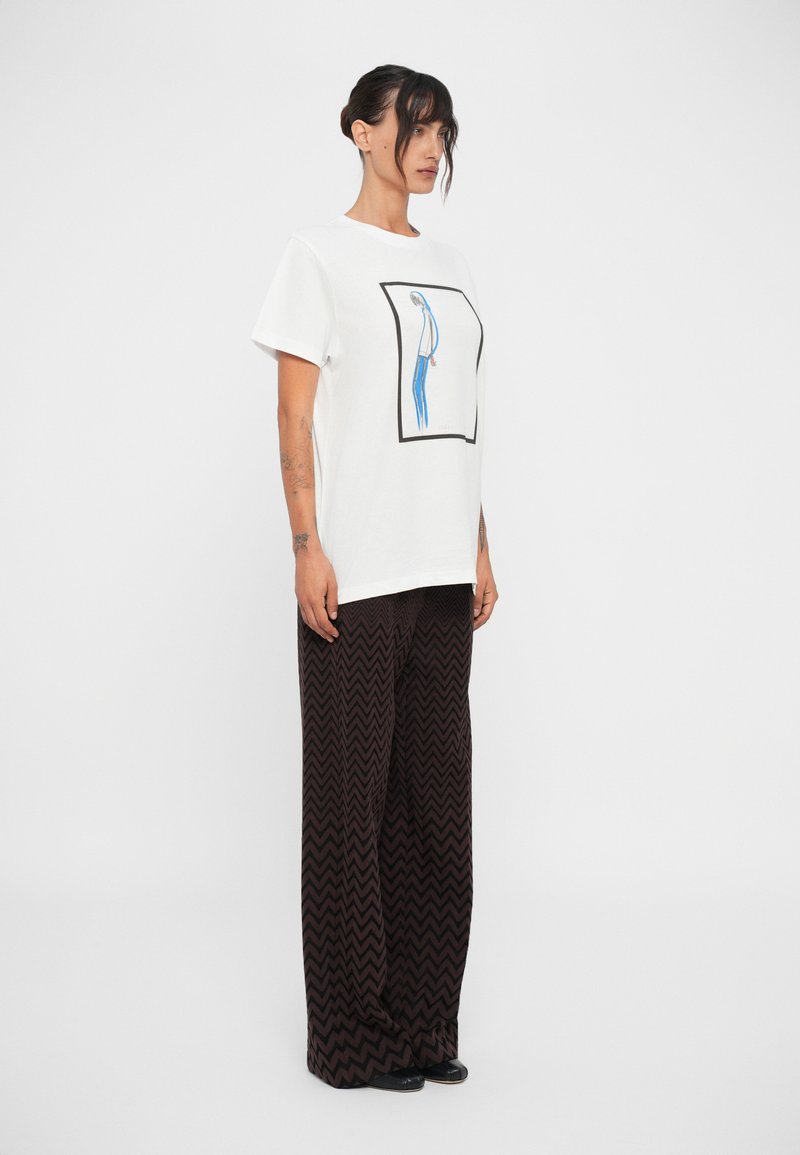 T-shirt bianco con una grafica blu e nera all'interno di un quadrato, abbinata a pantaloni a gamba larga marroni con motivi a zigzag. Tessuto morbido.