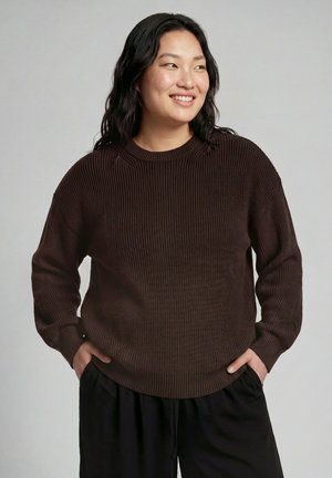 Femme aux longs cheveux noirs sourit en regardant vers la droite, portant un pull côtelé marron foncé et un pantalon noir, les mains dans les poches sur un fond uni.