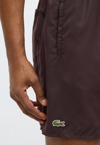 Shorts de bain marron foncé à texture lisse, avec des poches latérales et un logo crocodile vert brodé près de l'ourlet.