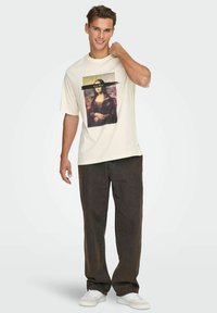 Cremefarvet bomulds-T-shirt med et grafisk print af Mona Lisa med sorte accenter. Kombineret med mørkebrune jeans og hvide sneakers.