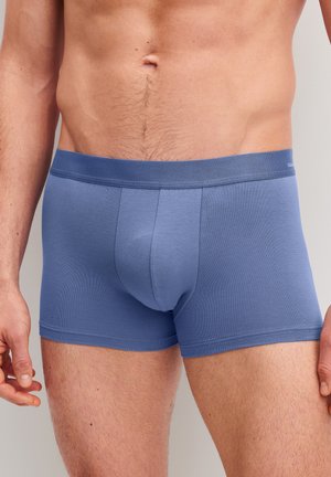 Mannelijke torso met nauwsluitende blauwe boxershorts, staand met ontspannen handen langs de zij tegen een effen achtergrond.