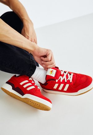 Personne portant des baskets rouges et blanches avec des lacets blancs, des chaussettes blanches et un pantalon noir, ajustant une chaussure en étant assise sur un sol blanc.