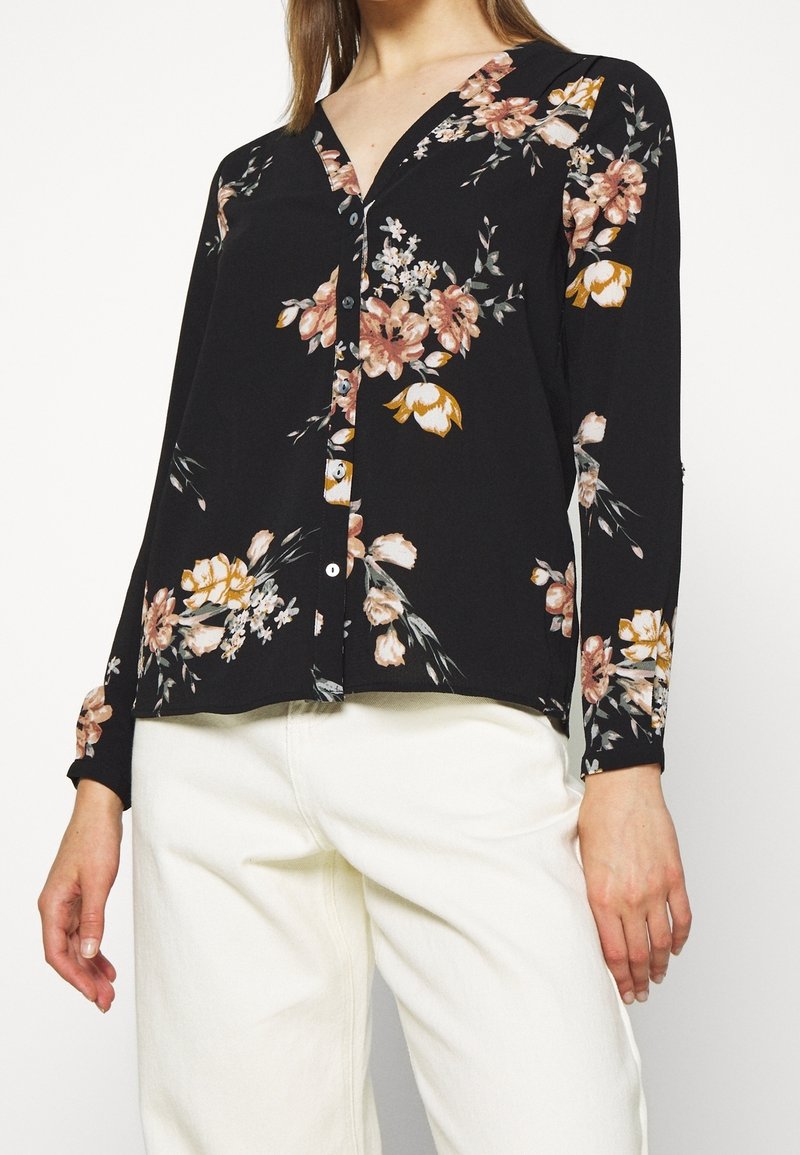 Femme portant un chemisier noir à manches longues avec des motifs floraux, boutonné sur le devant, associé à un pantalon blanc taille haute, debout devant un fond uni.