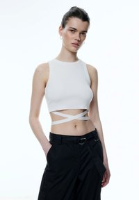 Witte geribde crop top met brede bandjes en gekruiste details bij de taille, gecombineerd met zwarte losse broek en een zilveren riemgesp.