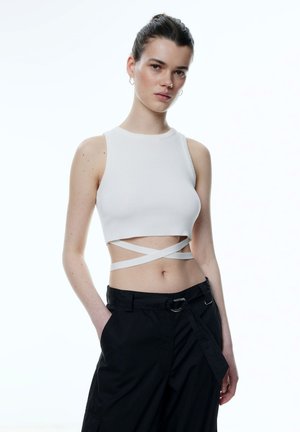 Witte geribde crop top met brede bandjes en gekruiste details bij de taille, gecombineerd met zwarte losse broek en een zilveren riemgesp.