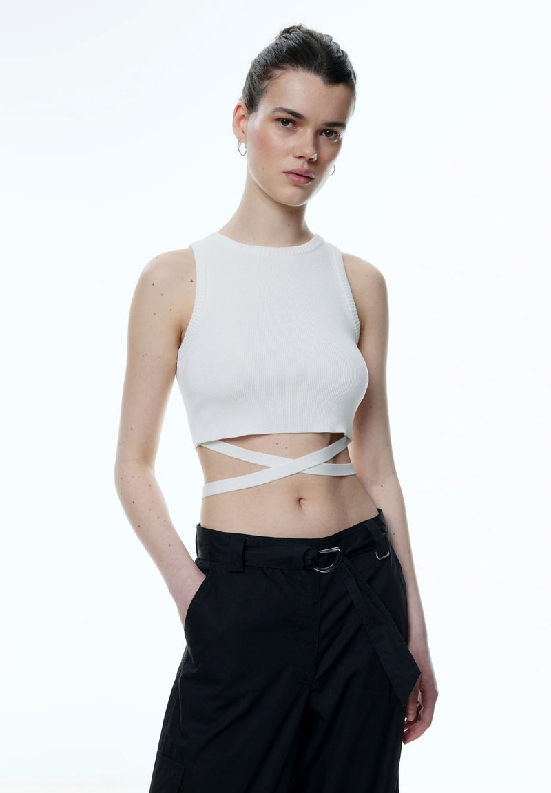 Witte geribde crop top met brede bandjes en gekruiste details bij de taille, gecombineerd met zwarte losse broek en een zilveren riemgesp.