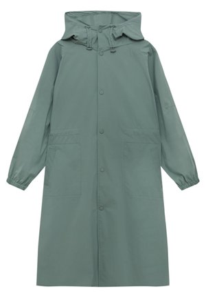 Persona che indossa un lungo cappotto verde salvia e pantaloni cargo, con un maglione a righe annodato in vita e scarpe da ginnastica bianche.