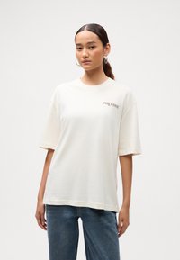 Joven mujer con aretes de aro, vistiendo una camiseta blanca de gran tamaño con el texto "PARK AVENUE" y jeans azules, frente a un fondo plano.
