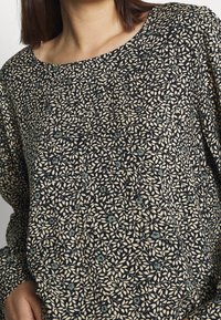 Schwarzes Oberteil mit floralem Muster in Creme und gedämpftem Grün. Rundhalsausschnitt, lange Ärmel, lockere Passform und weiche Textur.