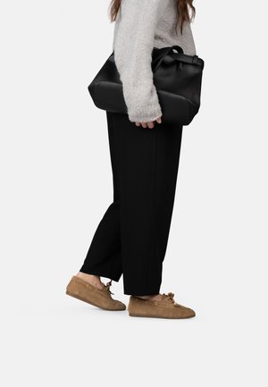 Vrouw draagt zwarte broek en beige mocassins, houdt een grote zwarte leren boodschappentas vast, en draagt een grijze, zachte trui.