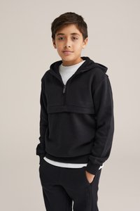 Svart zip-up hoodie med en texturerad nedre panel, bärs över en vit skjorta. Har en huva och framfickor, visas på en ung person.