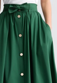 Gonna verde a pieghe con una larga fascia in vita, dettaglio a fiocco e bottoni dorati lungo la parte anteriore. Caratterizzata da tasche laterali e una consistenza liscia.